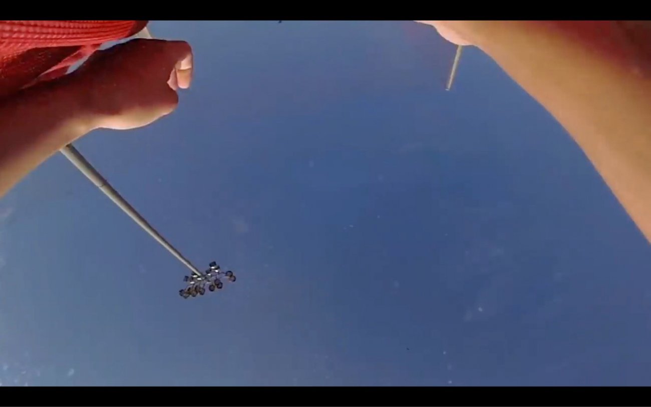 Image is a screenshot from a video. Image is looking from the ground up at the blue sky, stadium lights traverse the the lower center of the frame and out stretched arms frame either side of the image area.  