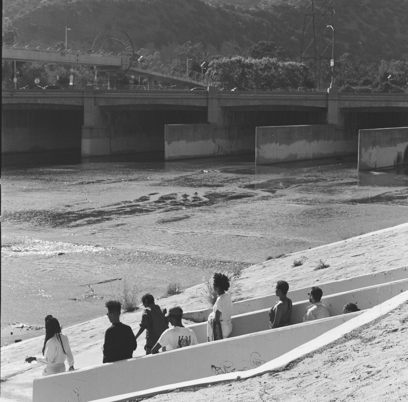 Black and white image still of a group of people seen from a distance looking out onto a reservoir. 