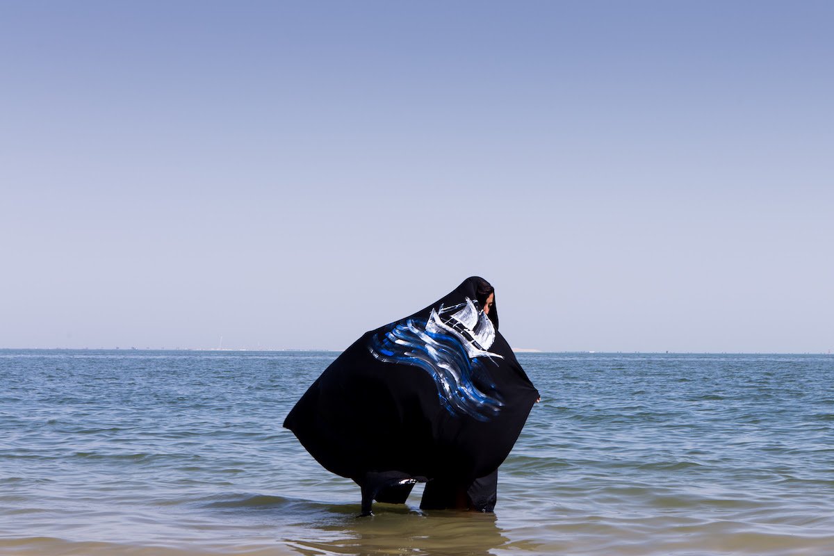 Photograph of person standing knee deep in water wearing a cloak with a ship painted on it.