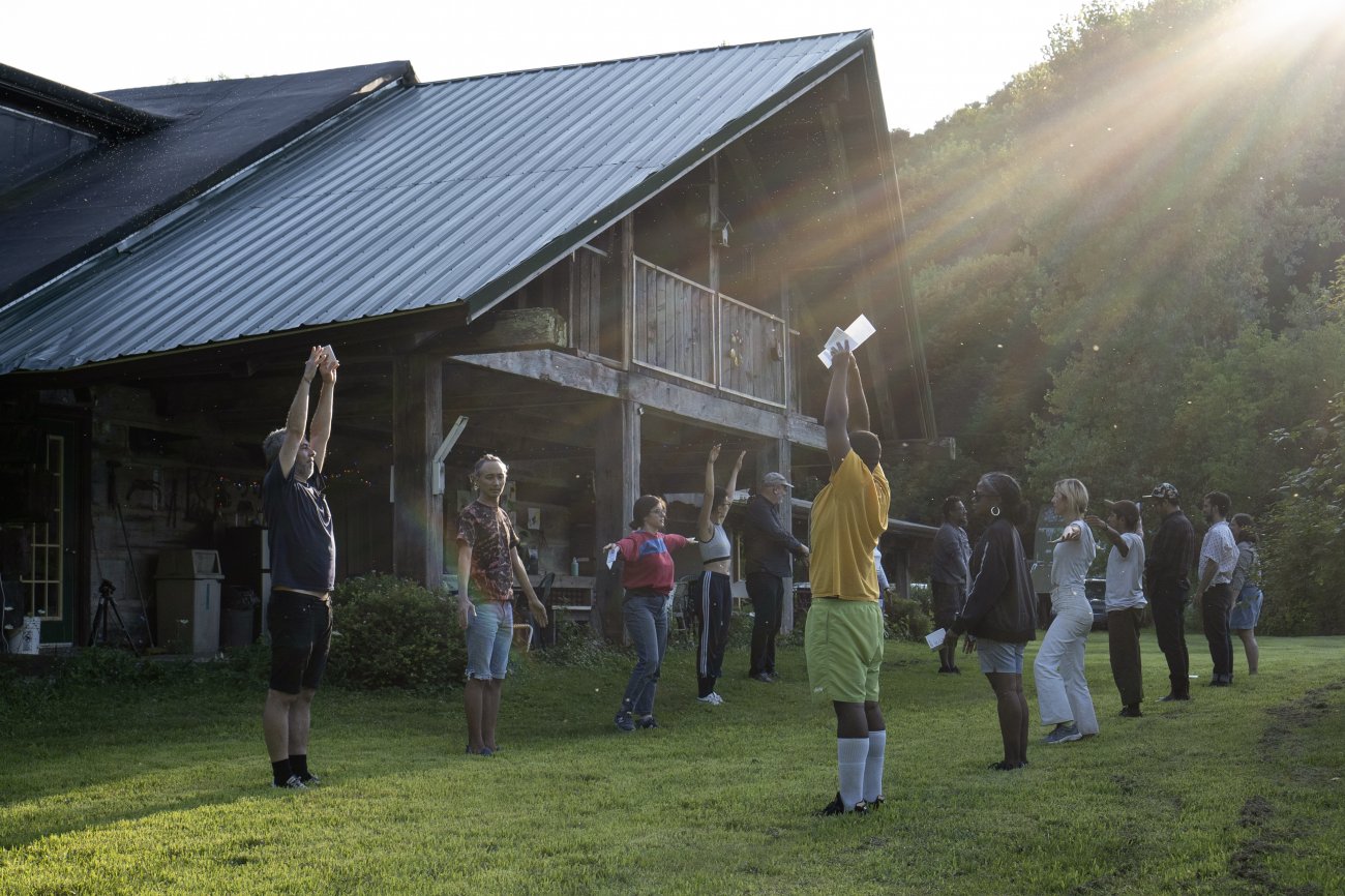 A group of people stretch their arms to the sky in the late afternoon sun rays. They are standing in two lines facing each other in front of the ACRE lodge.