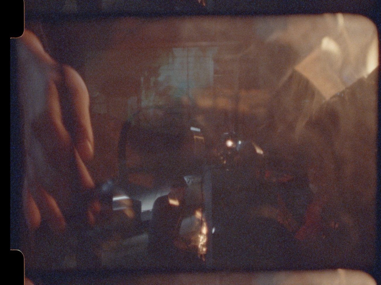 a still of a frame of a 8mm film shows a image with muliple exposure, hazy and brown with spots of light we see a hand entering the frame on the left manipulating the faint image of a machine.
