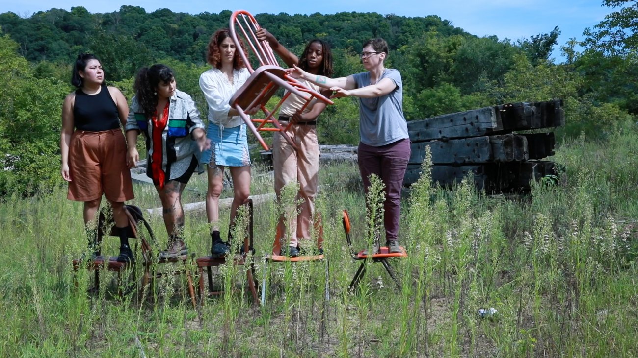 Five people standing in a row of chairs in the middle of a meadow. They are passing a red chair down the line.