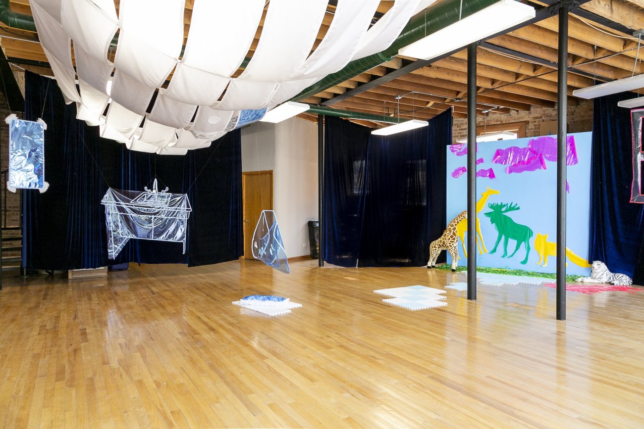 A wide view of the exhibition, featuring uniform, rectangular fabric panels swooping along the ceiling, clear vinyl objects suspended from the ceiling (two abstract and one resembling a sink), modular foam floor tiles, and a backdrop featuring two-dimensional animal cut outs, and stuffed animals flanking each side of the backdrop.
