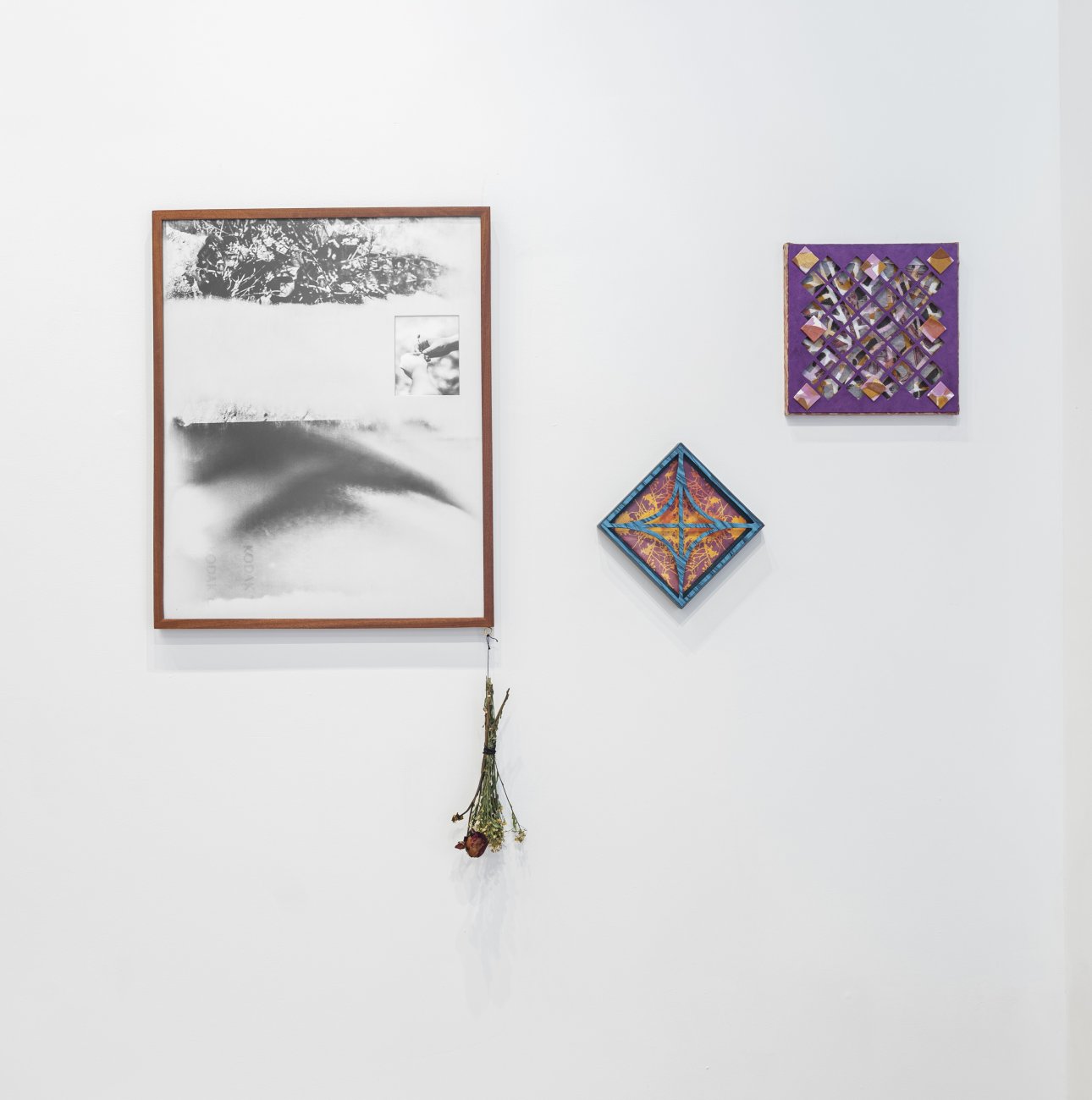 Black and white abstract image framed in a thin wood frame with a dead bouquet of flowers hanging off the bottom right corner. To the work's right are two small sculptural collages with paper cutouts.