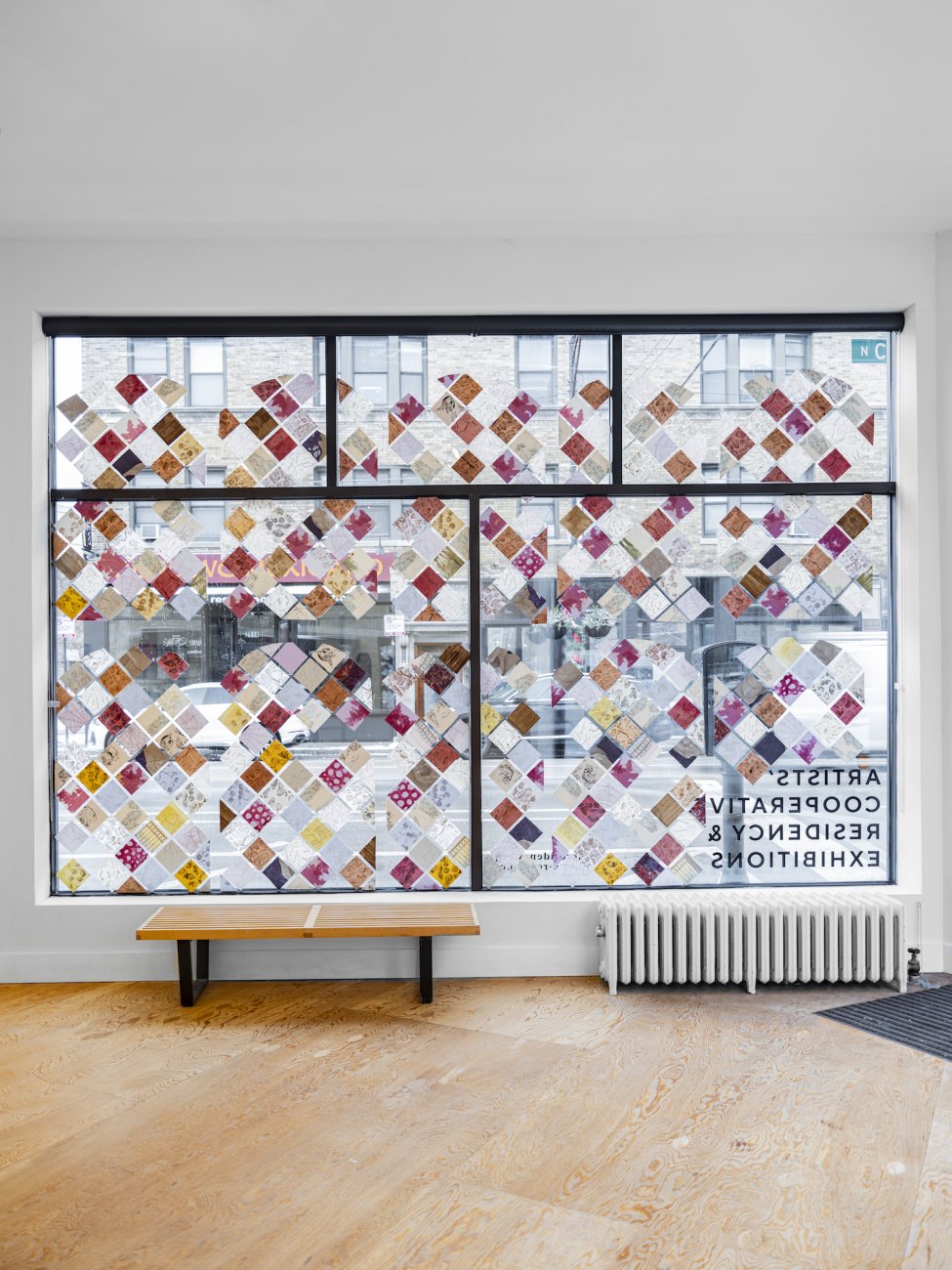 The window installation in ACRE's storefront window. Small diamond-shaped papers of various colors and patters are adhered to the window in an orderly linear pattern.