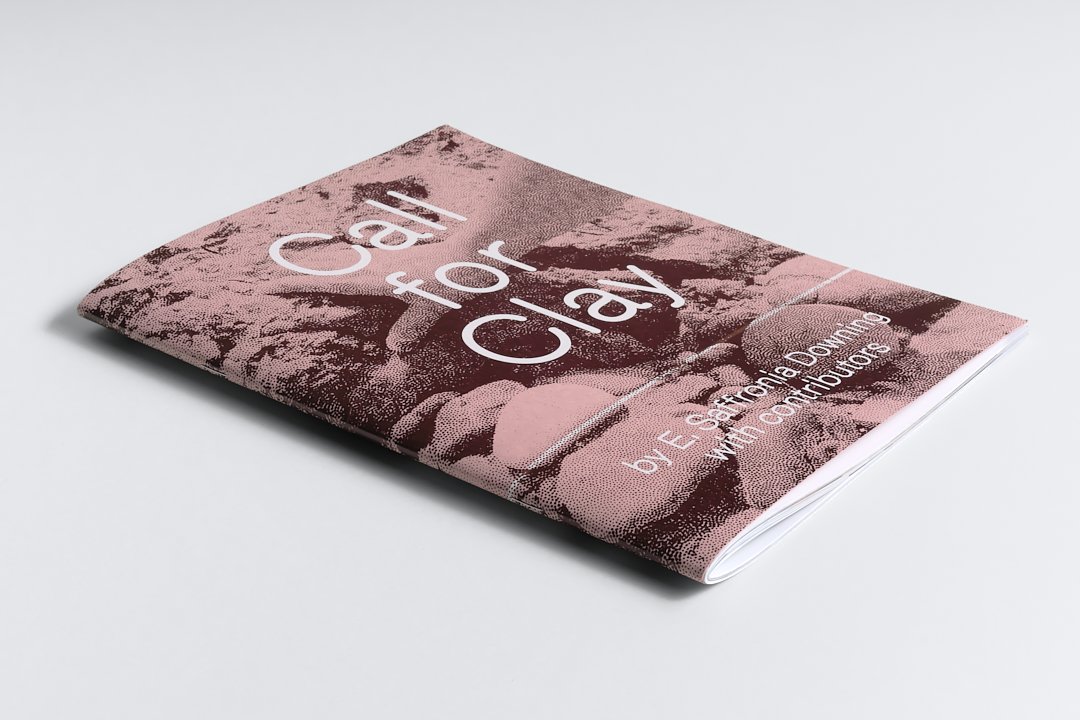A copy of the softcover "Call for Clay" publication lays closed on a table. The cover is a sepia toned image of a hand reaching towards clay and rocks overlaid with white text reading "Call for Clay by E. Saffronia Downing with contributors". 