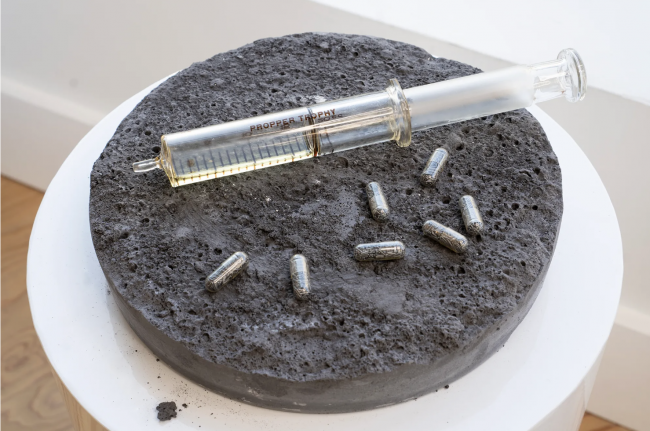  A glass syringe text reading “Propper Trophy” rests on a craterous graphite-like disc alongside seven pill capsules filled with copper. There is liquid in the syringe but it is almost empty.