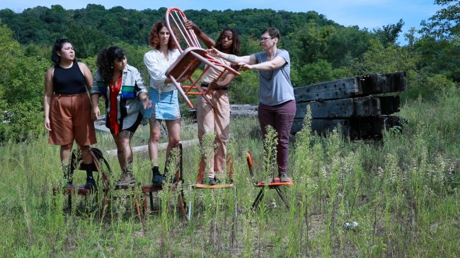 Five people standing in a row of chairs in the middle of a meadow. They are passing a red chair down the line.