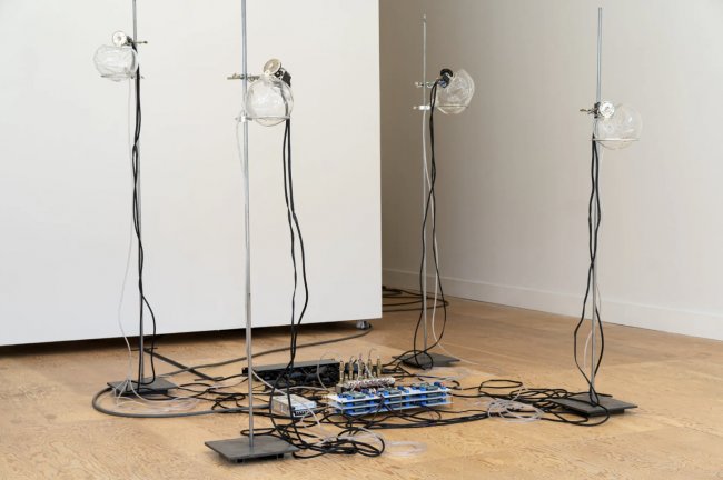 Four blown glass orbs resting on metal stands. Attached to all four orbs, chords and wires collect in a pile on the floor in the center of the sculptures.
